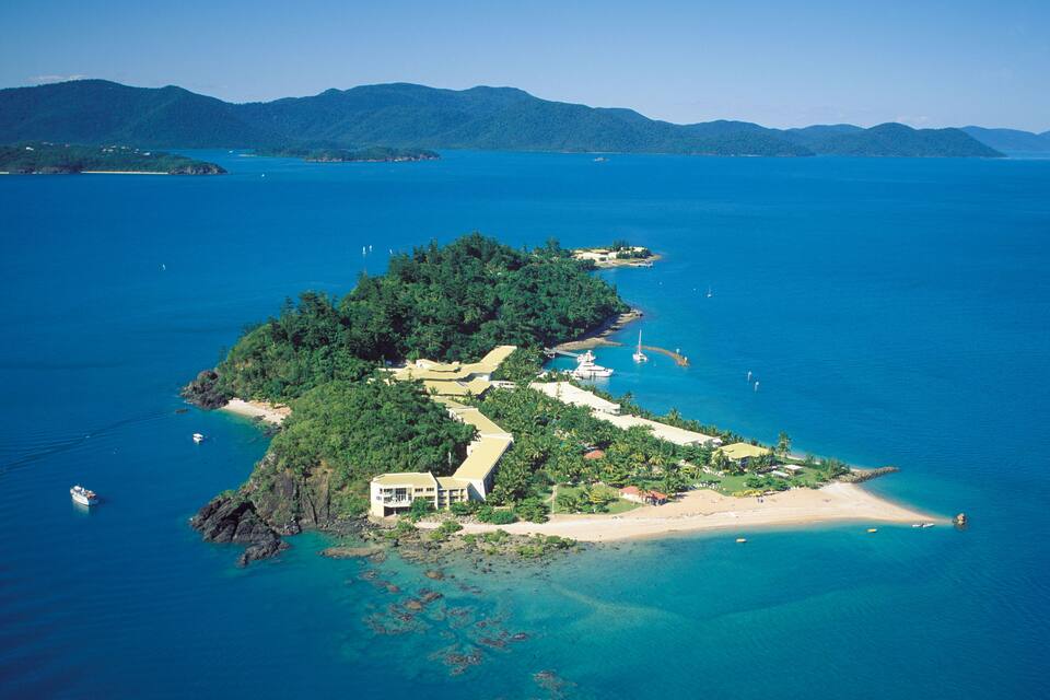 An aerial view of a tropical island with a large building, clear blue waters, and distant mountains.