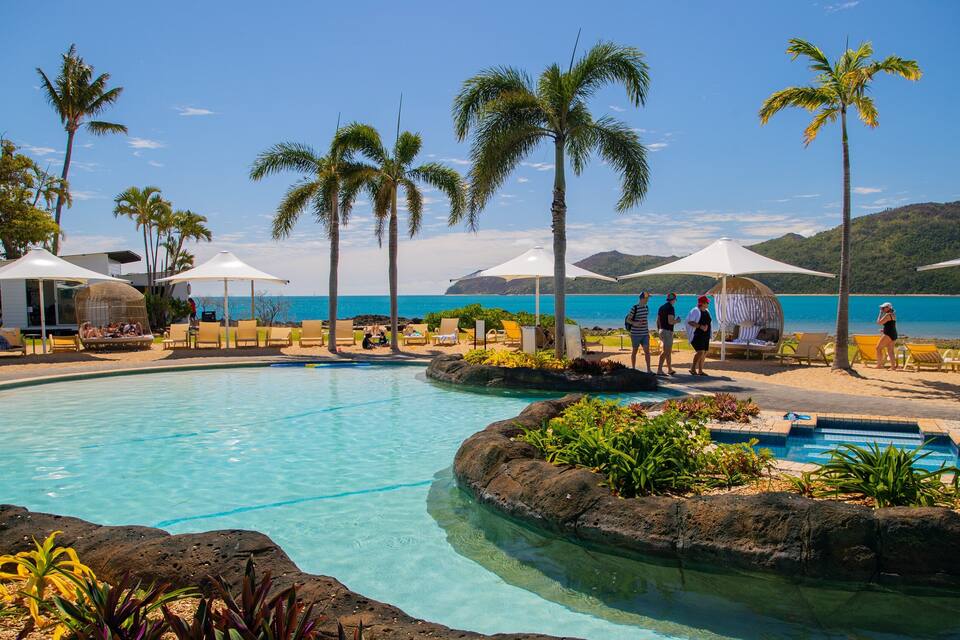 A poolside area with lounge chairs and umbrellas, palm trees, and a view of the ocean.