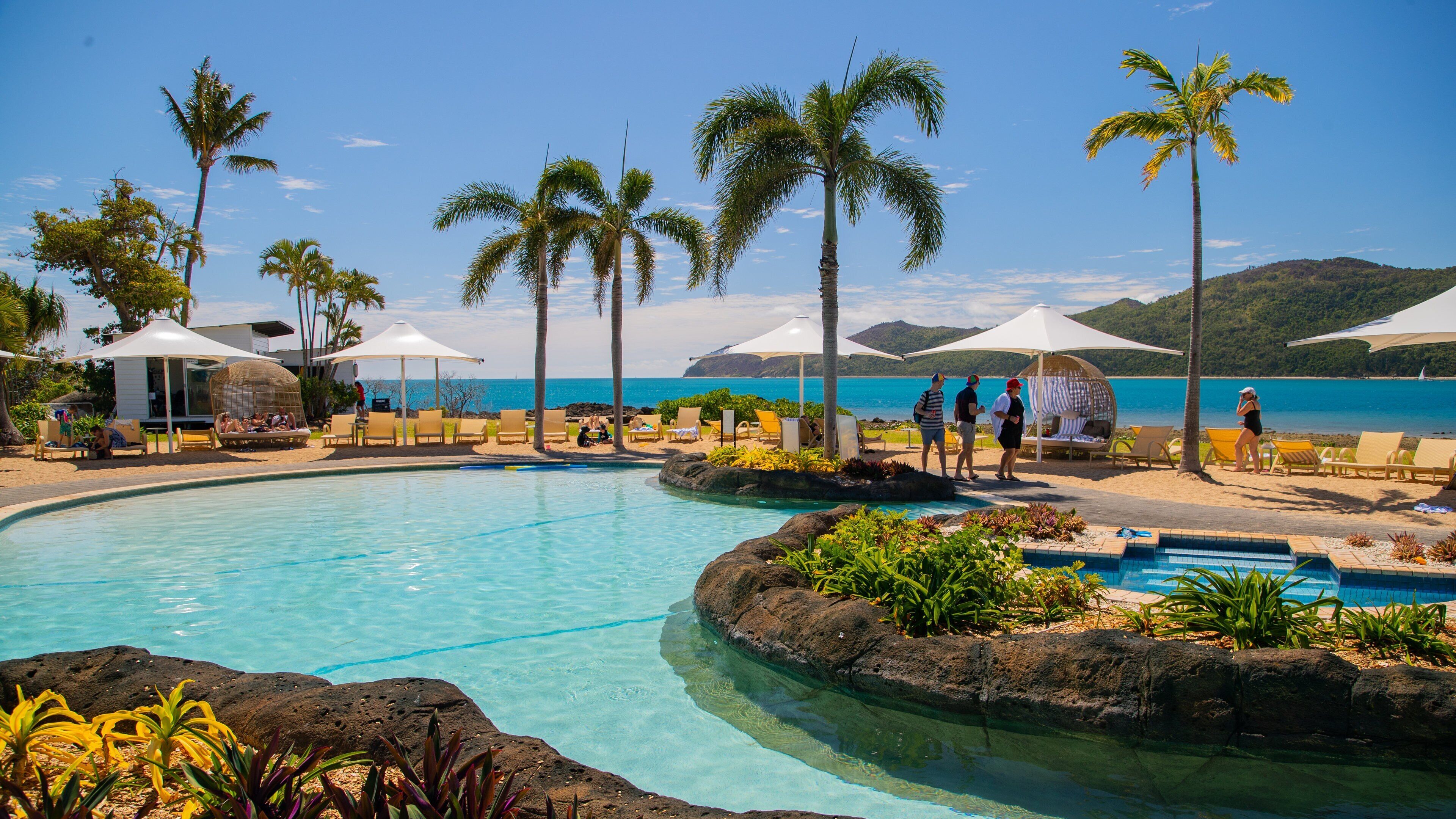 A poolside area with lounge chairs and umbrellas, palm trees, and a view of the ocean.