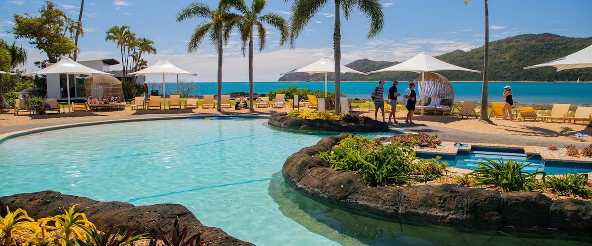 A poolside area with lounge chairs and umbrellas, palm trees, and a view of the ocean.