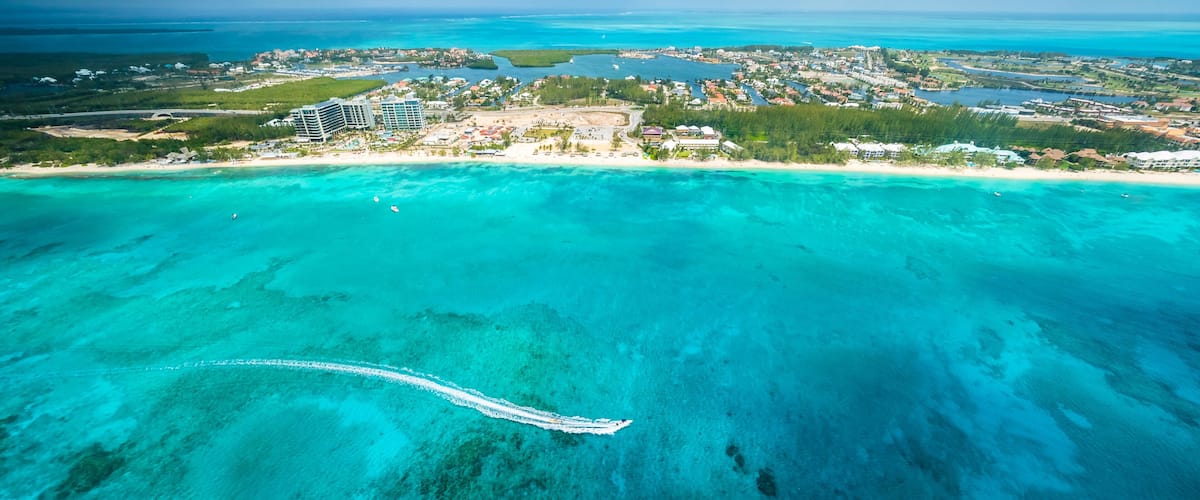 An aerial view of a coastal area with clear turquoise waters, a white boat trail, and a distant town with buildings and greenery.