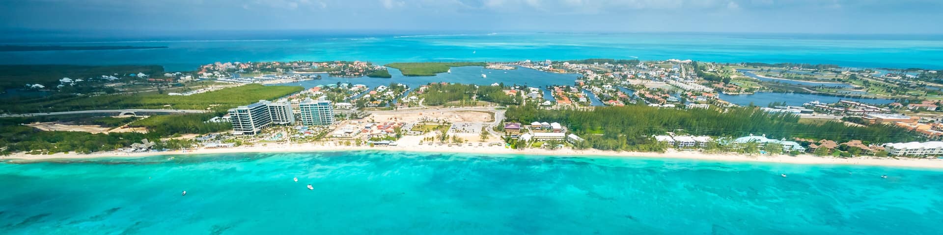 An aerial view of a coastal area with clear turquoise waters, a white boat trail, and a distant town with buildings and greenery.
