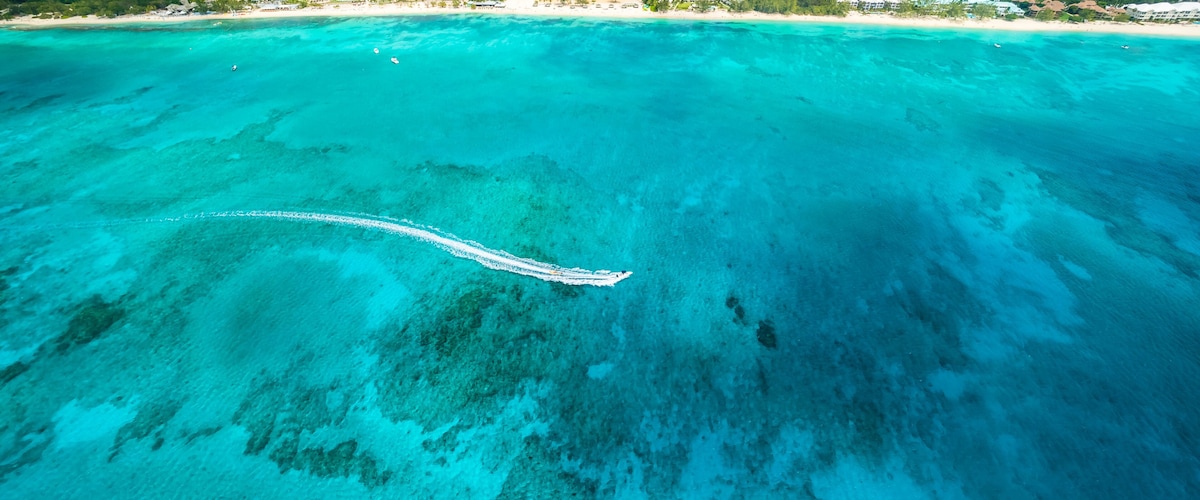 An aerial view of a coastal area with clear turquoise waters, a white boat trail, and a distant town with buildings and greenery.