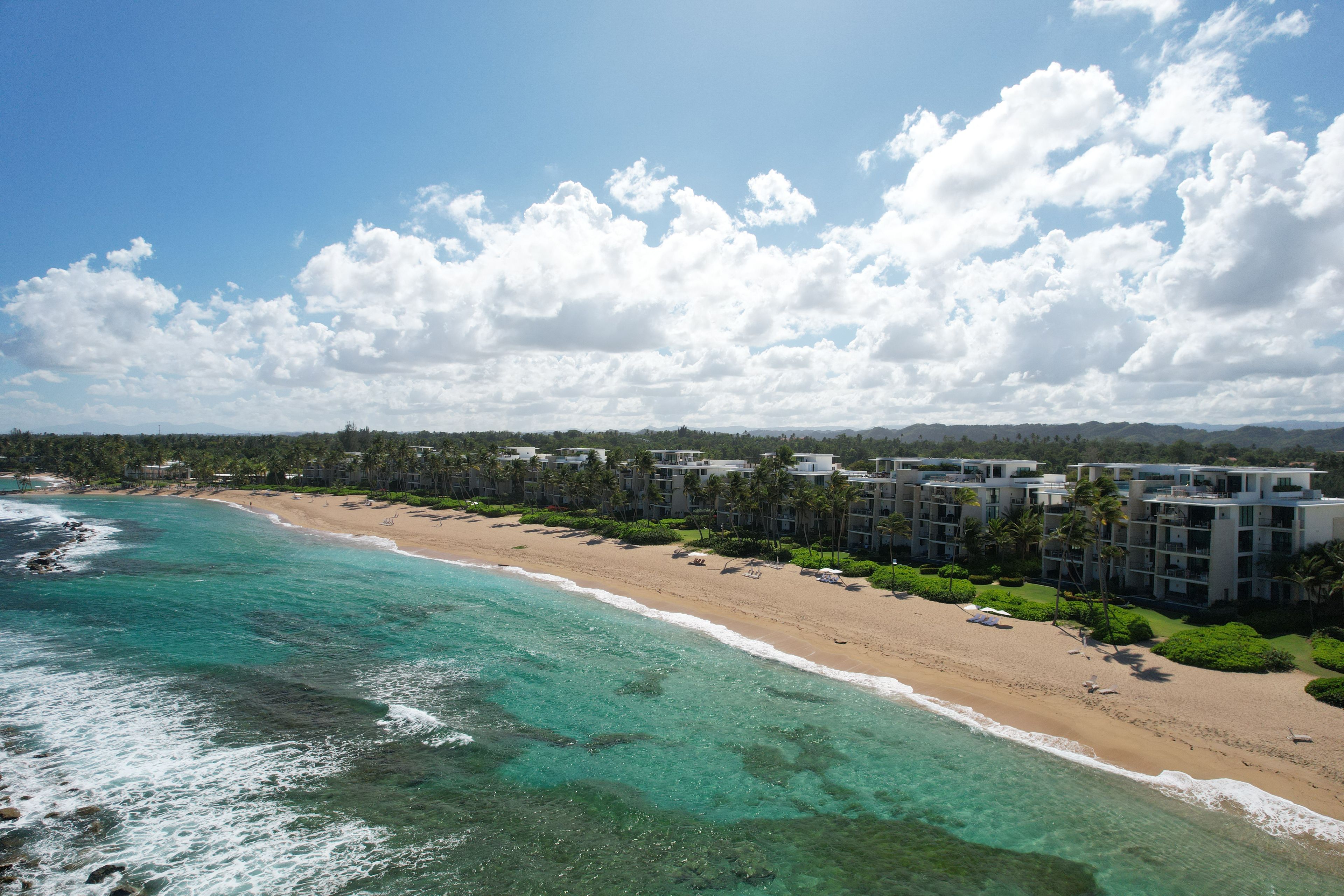 A beach with clear turquoise waters, a sandy shore, and a row of modern buildings with palm trees.