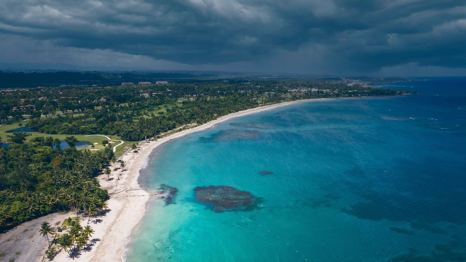A coastal landscape with a beach, clear turquoise waters, and a backdrop of cloudy skies.