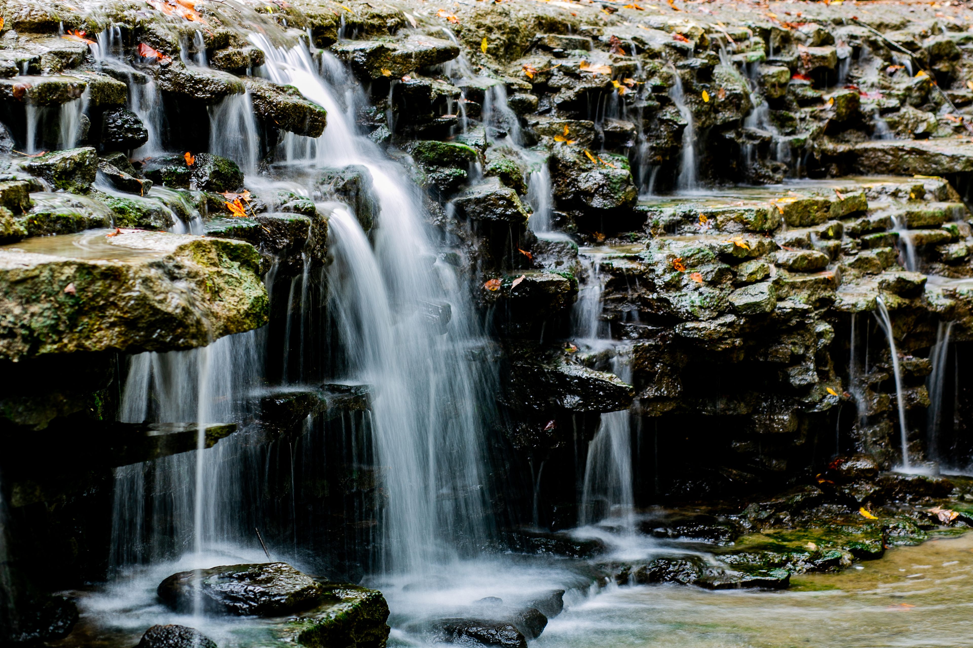 View of a tiered waterfall cascading over dark, mossy rocks, adorned with scattered autumn leaves in Sharonville, Ohio, United States.