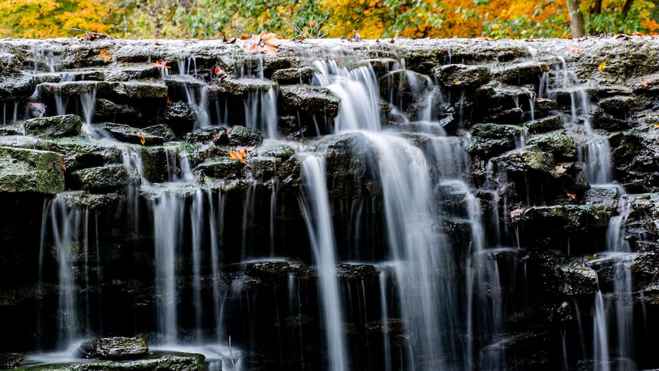 A cascading waterfall over a rocky ledge with autumn foliage in the background.