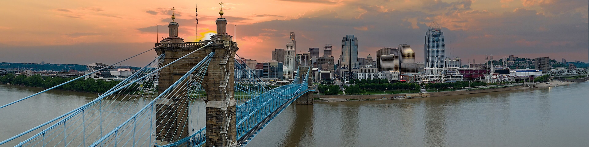 A blue suspension bridge over a river with a city skyline in the background.