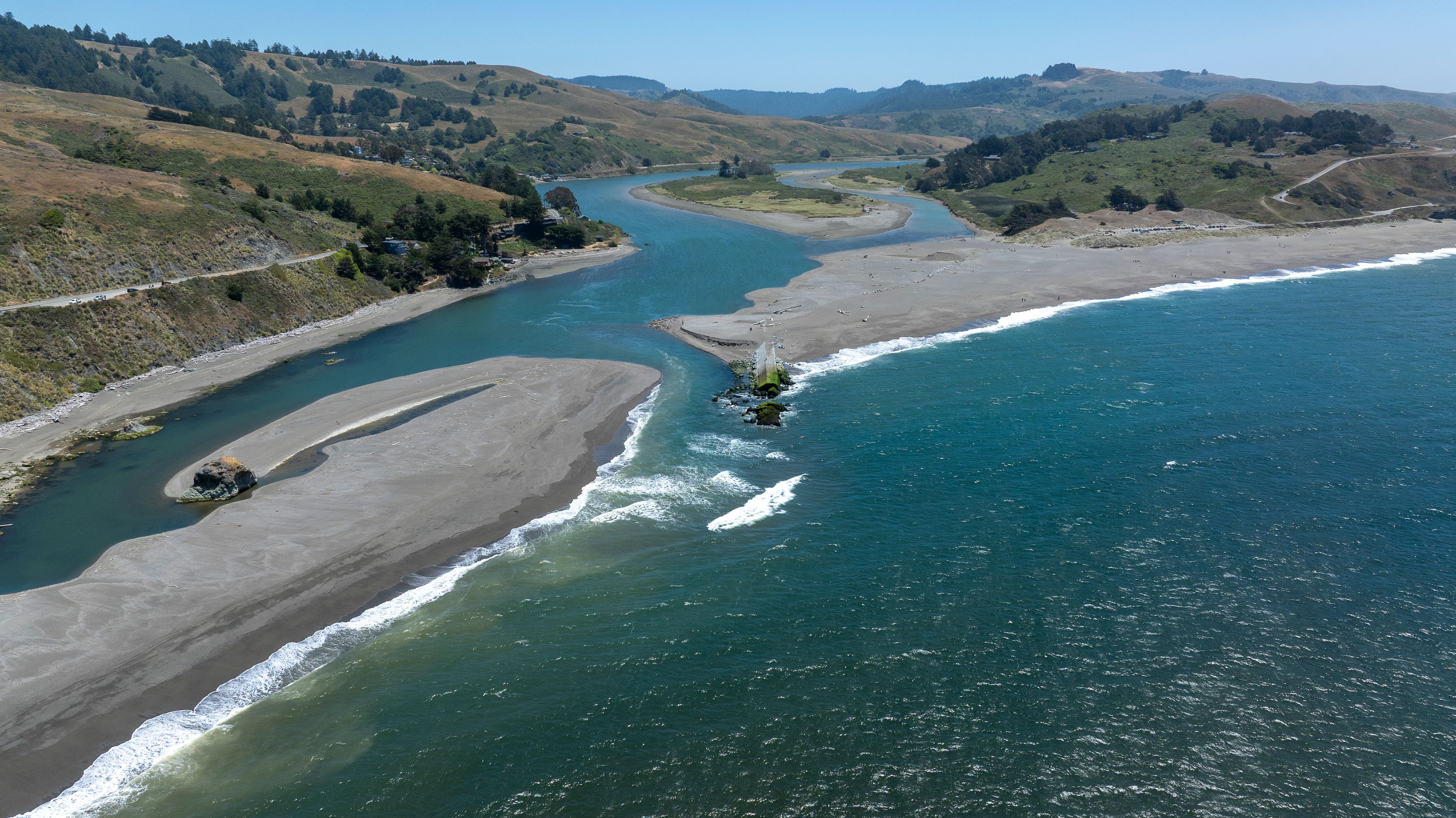 A river flowing through a valley with hills on either side, a beach, and a clear sky.