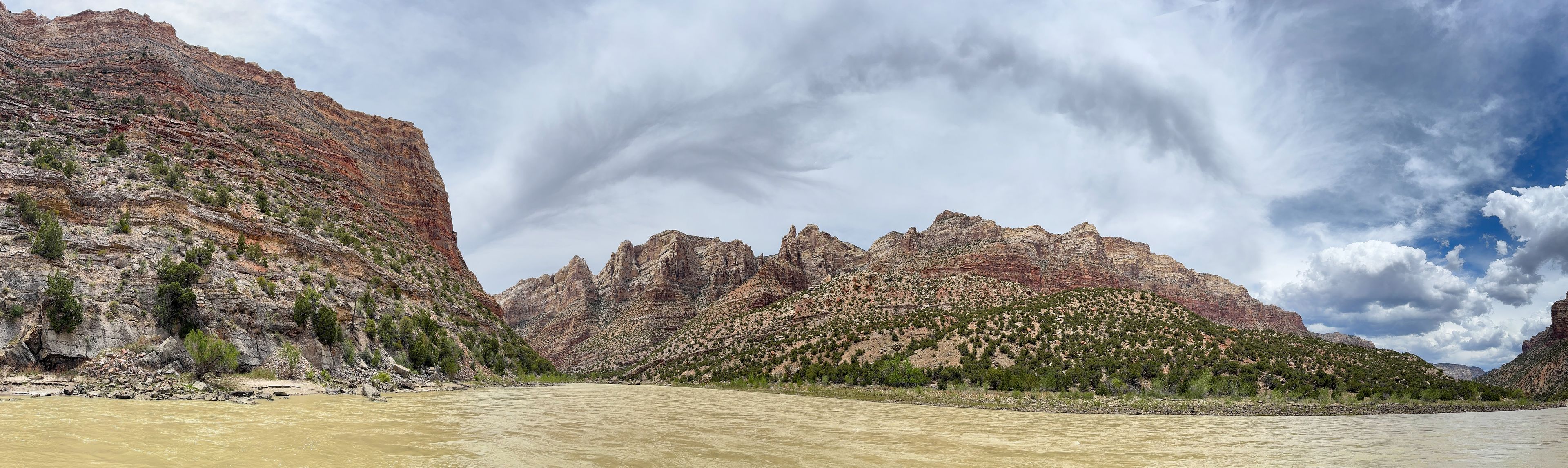 View of the Yampa River flowing through a dramatic canyon with rugged red rock cliffs, sparse vegetation, and a cloudy sky in Moffat County, Colorado, United States.