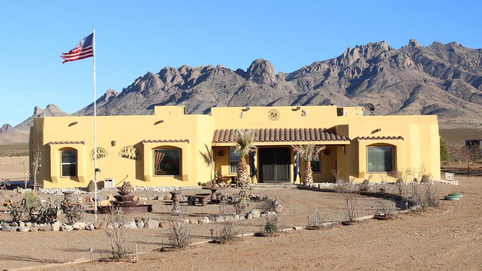 A yellow building with a flag, surrounded by desert vegetation and mountains.
