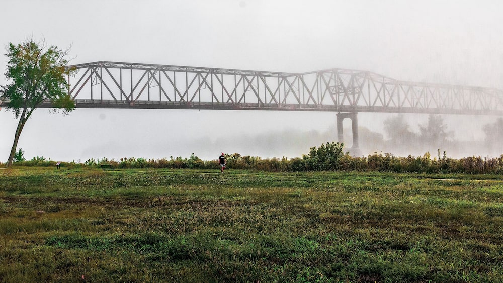 Eine Brücke über ein Gewässer, umgeben von einer Wiese mit einem einzelnen Baum und einer Person, die unterwegs ist.