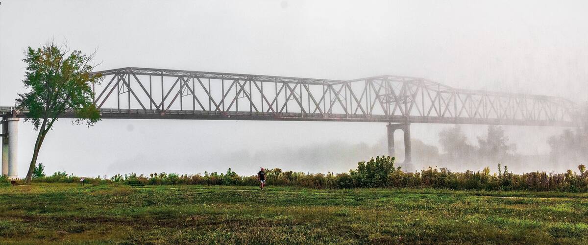 A bridge over a body of water, surrounded by a grassy field with a single tree and a person walking.