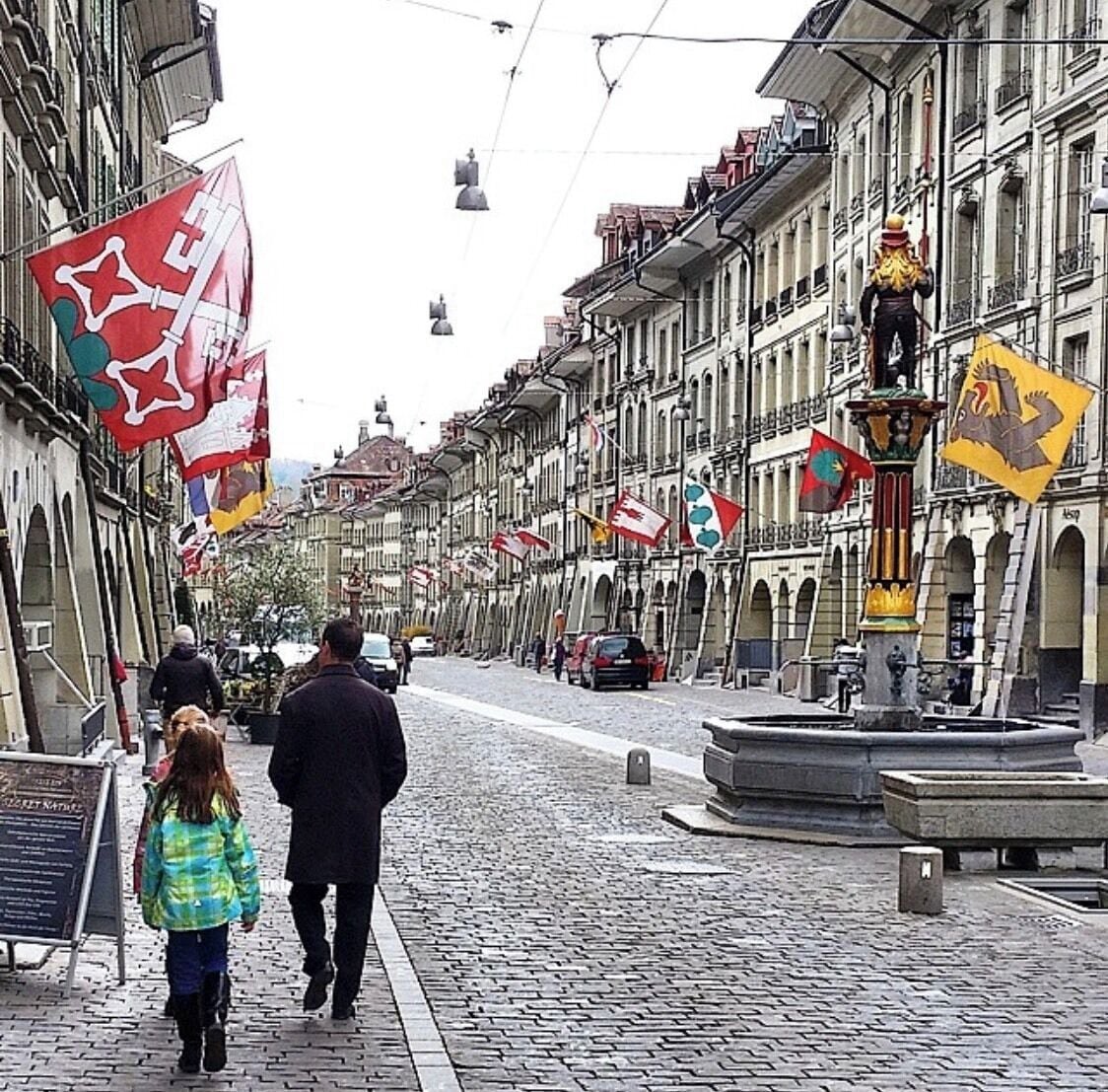 A cobblestone street lined with historic buildings, flags, and a statue.