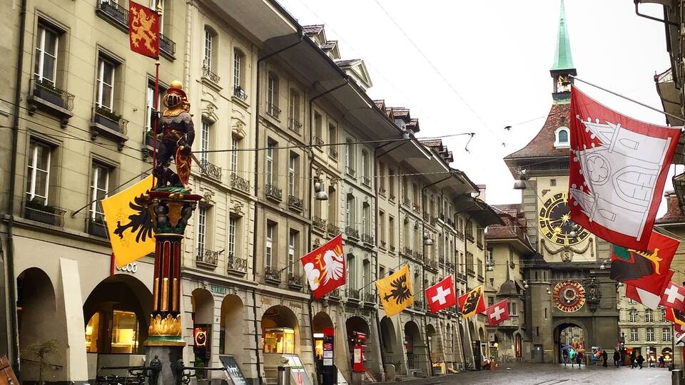 A cobblestone street lined with historic buildings, flags, and a fountain.