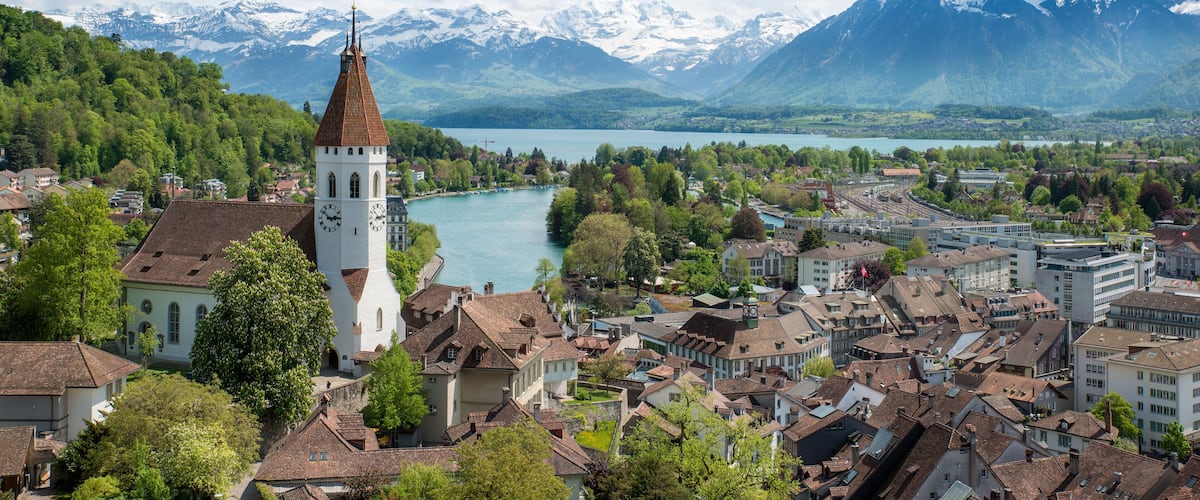 A town with a prominent church tower, a river, and snow-capped mountains in the background.