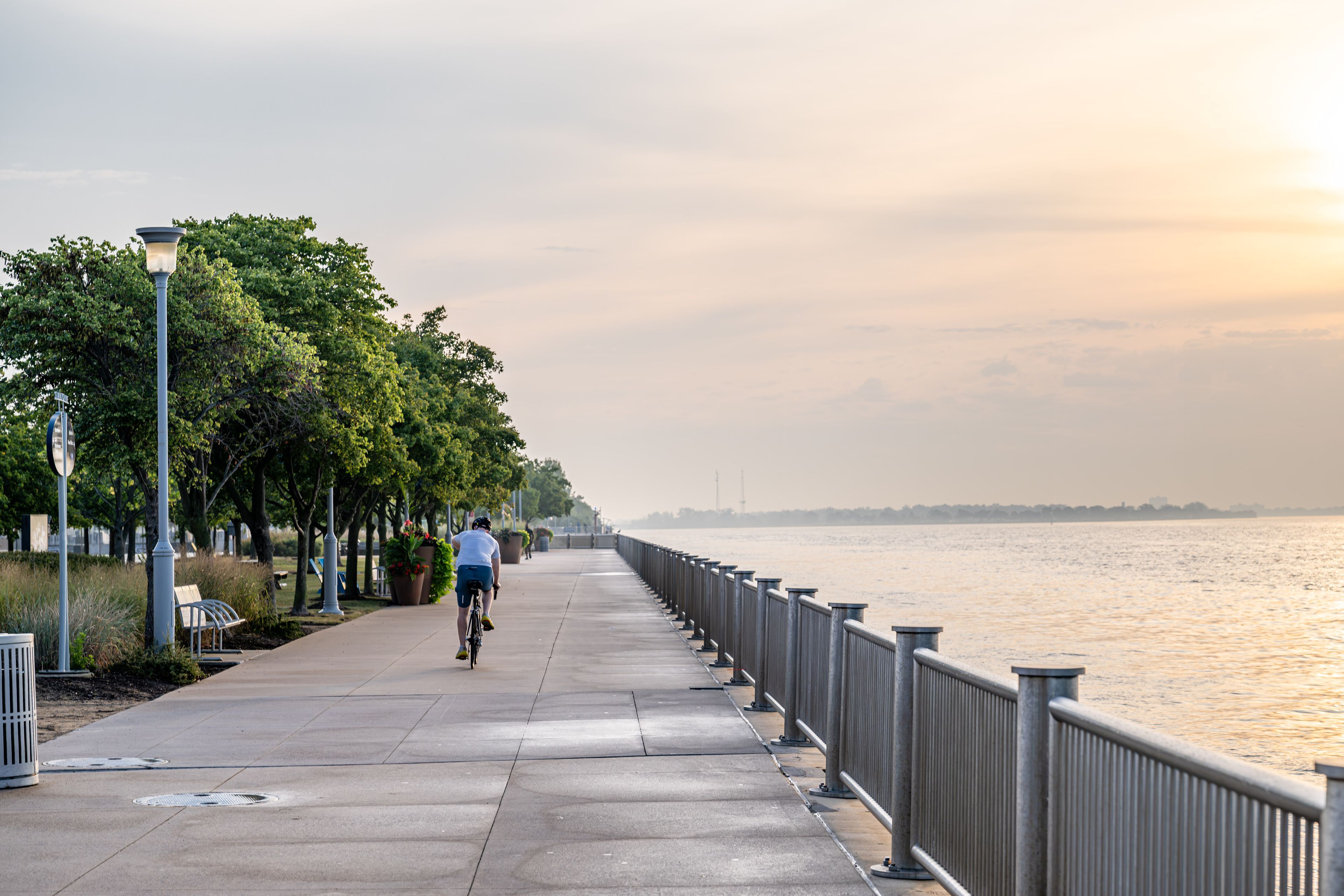 A waterfront promenade with a cyclist, trees, benches, and a railing along the path.