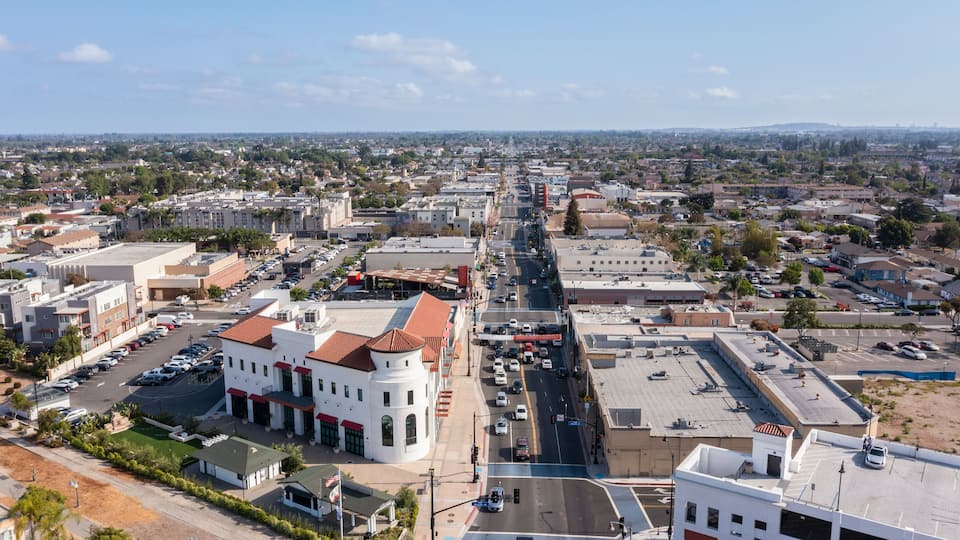 A cityscape with a prominent white building with a red roof in the foreground, a wide street, and a distant view of a city skyline.