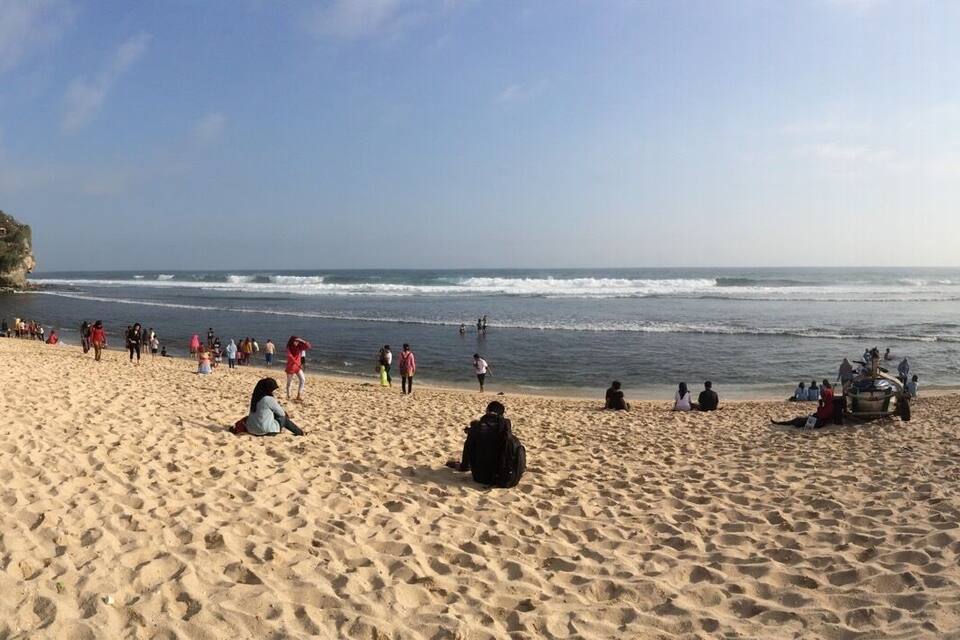 Uma praia arenosa com pessoas sentadas e caminhando pela margem, um afloramento rochoso ao fundo e um céu claro.