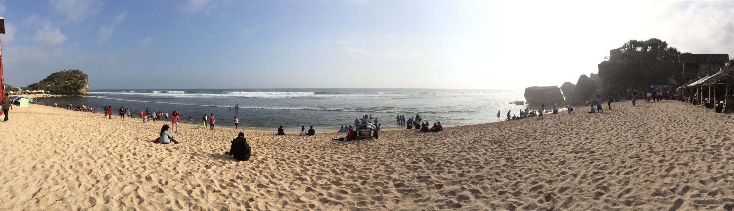 A sandy beach with people sitting and walking along the shore, a rocky outcrop in the background, and a clear sky.