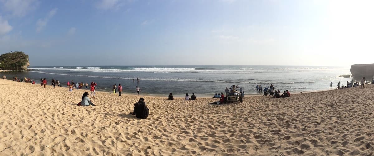 A sandy beach with people sitting and walking along the shore, a rocky outcrop in the background, and a clear sky.