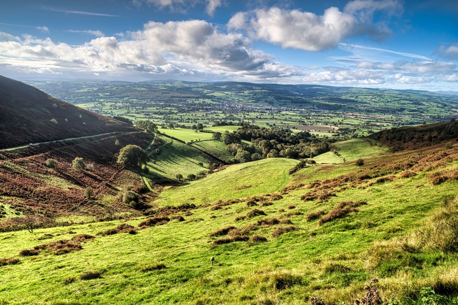 Rolling hills, a winding road, and a distant village under a partly cloudy sky.