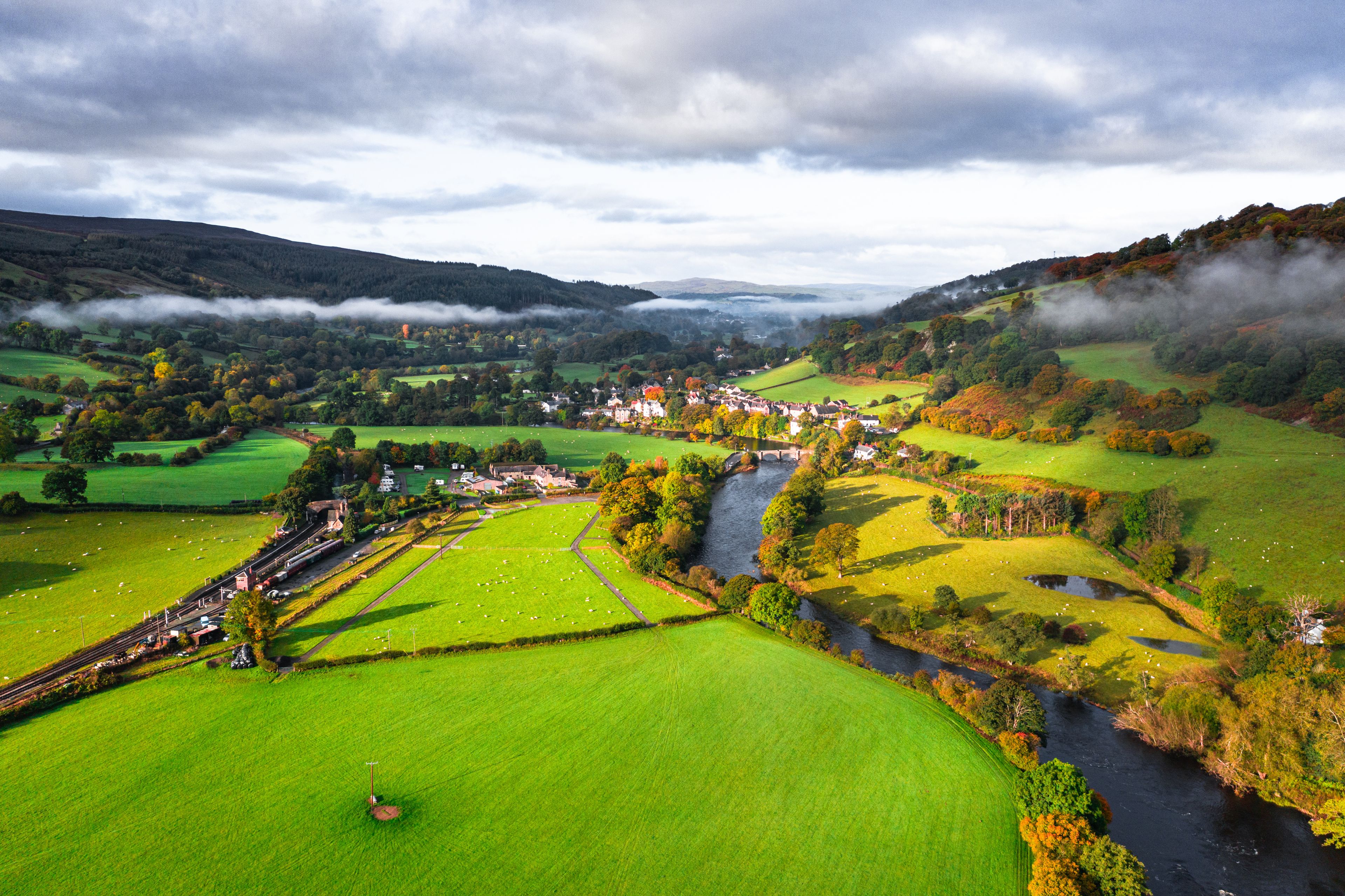 Paisagem rural com rio, campos verdes, uma pequena aldeia e colinas ondulantes.