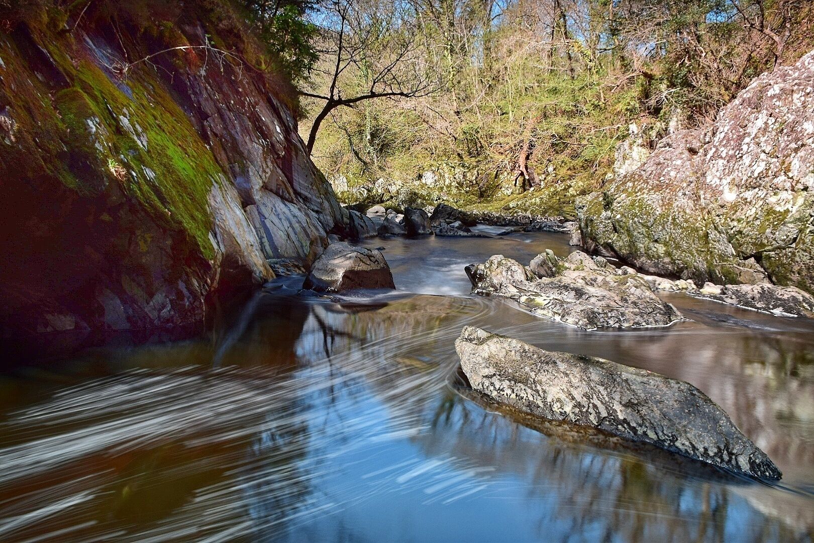 A river flowing through a rocky landscape with moss-covered rocks and trees in the background.