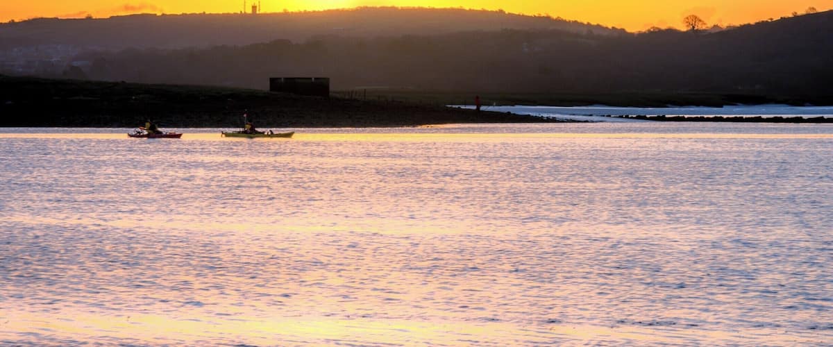 A sunset over a calm body of water with boats and a distant hill.