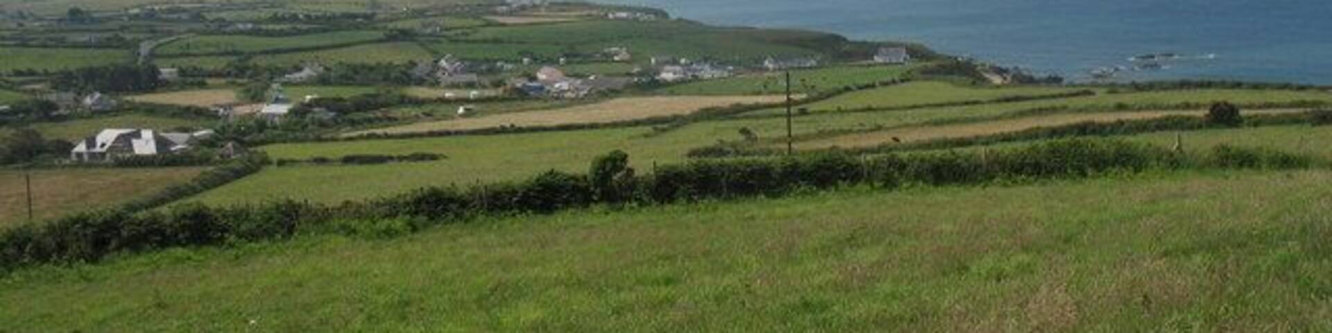 A coastal landscape with rolling hills, scattered houses, and a distant view of the sea and a mountain.