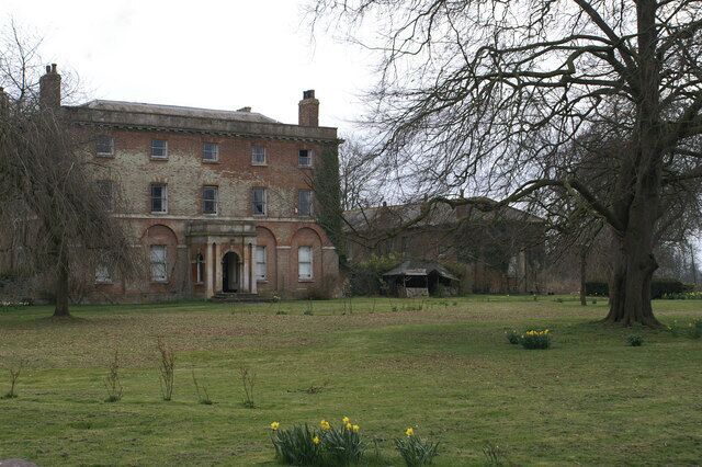 A large brick building with a central entrance, surrounded by a grassy field and trees.
