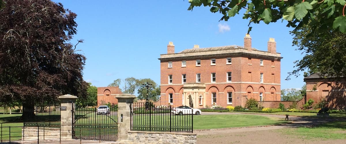 A large brick building with a central tower, surrounded by a well-maintained lawn and a stone bridge.