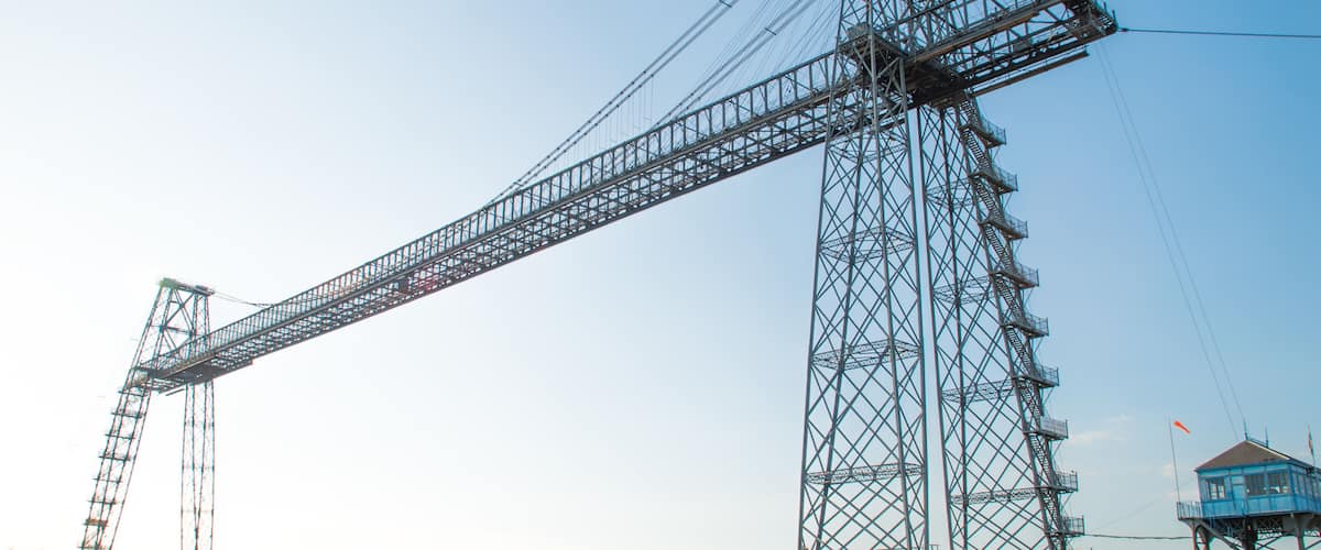Uma grande ponte treliçada de aço atravessa um rio, com uma torre de controle e edifícios residenciais ao fundo.