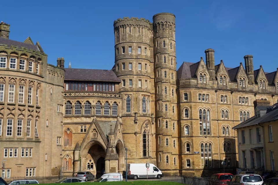 A historic building with a large arched entrance, tall cylindrical towers, and a mix of Gothic and Tudor architectural styles.
