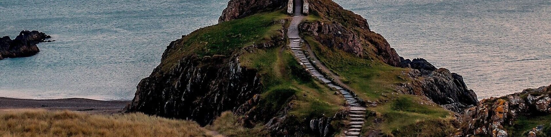 A lighthouse on a rocky outcrop with a path leading to it, overlooking the sea at sunset.
