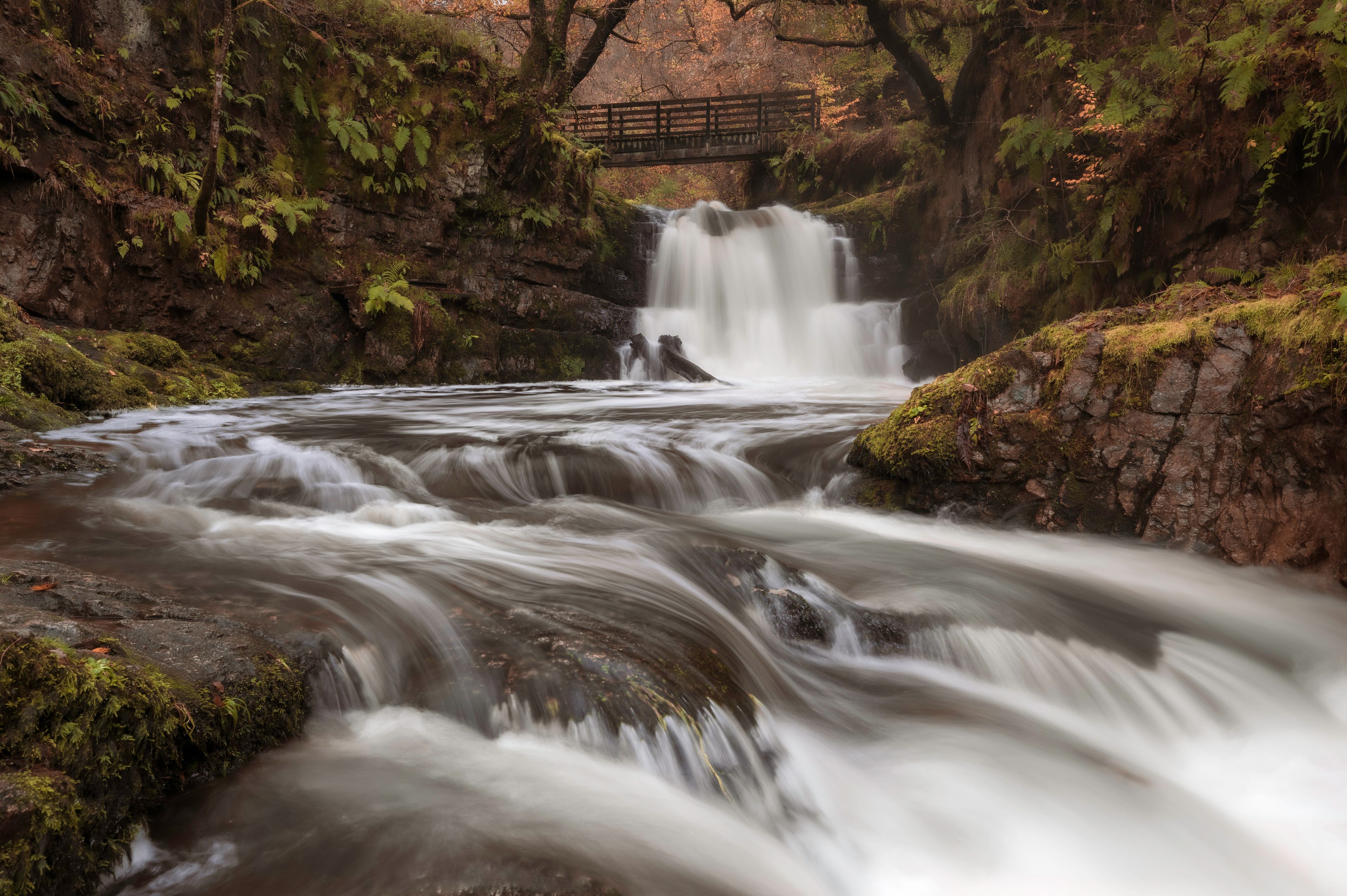A waterfall cascading over rocks in a forested area with a wooden bridge in the background.