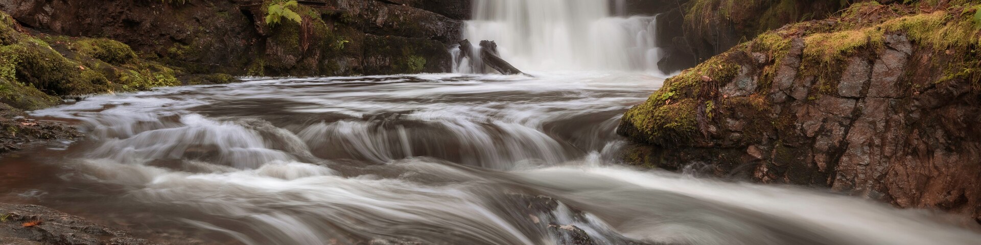 A waterfall cascading over rocks in a forested area with a wooden bridge in the background.