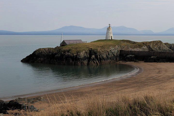 Un faro en una isla rocosa con una playa en primer plano y montañas al fondo.