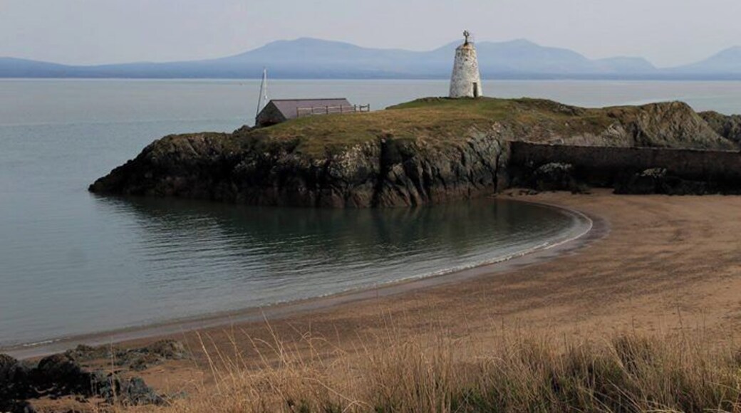 Un faro en una isla rocosa con una playa en primer plano y montañas al fondo.