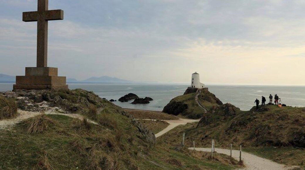 Una gran cruz de piedra, un faro y un camino costero con gente a lo lejos.