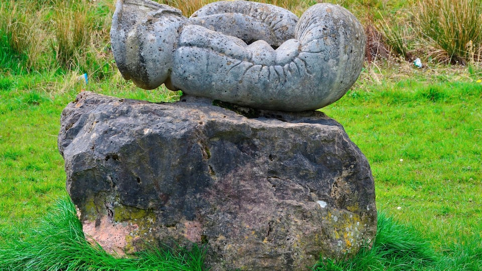 Sculptor Robert Kennedy.
Six separate small upright works set on stone bases; two boxing gloves, two dolls, cherub and a skull. Information from Festival Park: 'The works were carved away from site during 1991. They are made to operate as conventional garden ornamentation, within a constructed garden. The boxing gloves are there to depict some of their associated aspects, such as maleness and competition. The dolls depict something of how an image is idealised for mass consumption, becoming a distortion of the human figure. Both the gloves and the dolls can be seen as gendering devices through role play. The cherub and skull are life and death charms. Each of these suggests ways in which we adopt roles in society. What we really are is never as clear cut as what these images suggest.'
Borderline was supported by Garden Festival Wales and Chapter Arts Centre. 'The sculptor, Robert Kennedy, felt that the Garden Festival was a huge, themed social event in itself. He wanted to create a themed garden within it, something social, commenting to some extent on the diversity of culture within real society.'
A number of sculptures were commissioned for the Garden Festival of Wales in 1991/2 and some works remain on site.