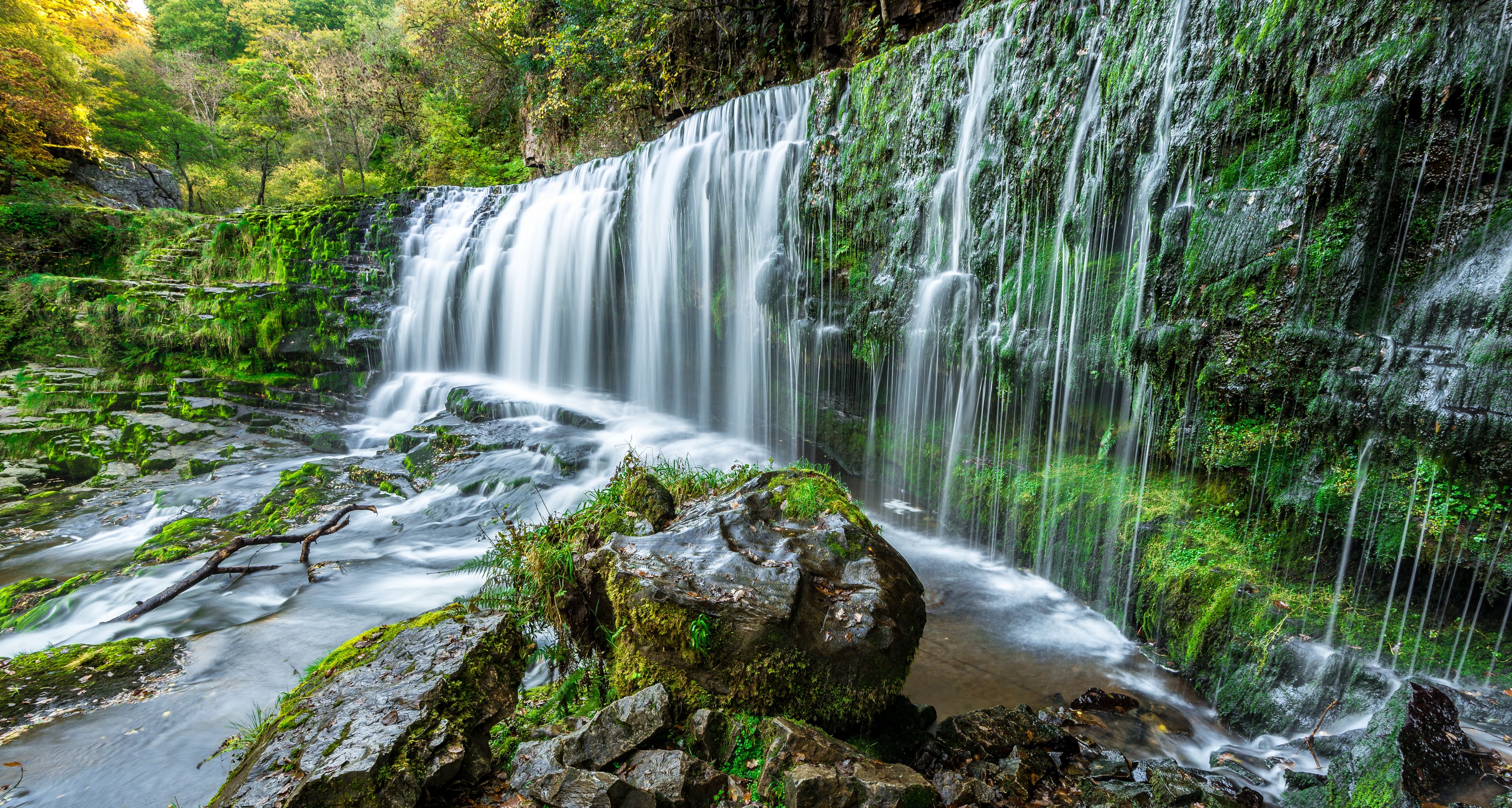 Uma cascata despencando sobre rochas cobertas de musgo em uma área arborizada.
