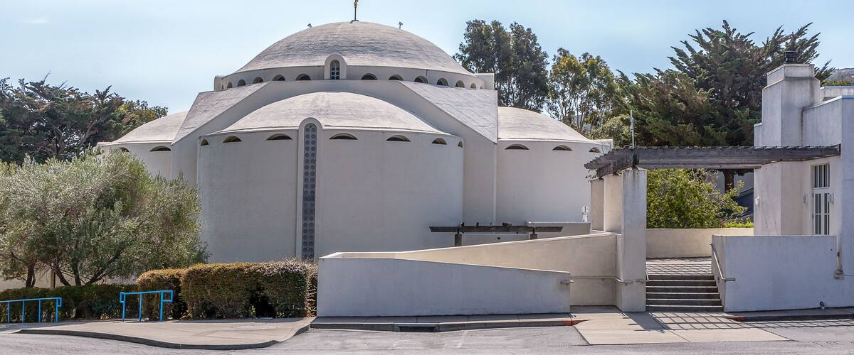 A white building with a dome and arched windows, surrounded by trees and a clear sky.