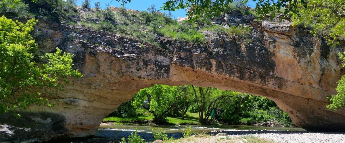 A natural stone bridge over a river with surrounding greenery and a clear blue sky.