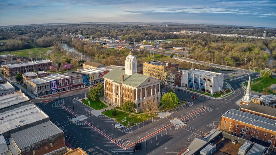 An aerial view of a town with a prominent building with a clock tower, a river, and surrounding greenery.