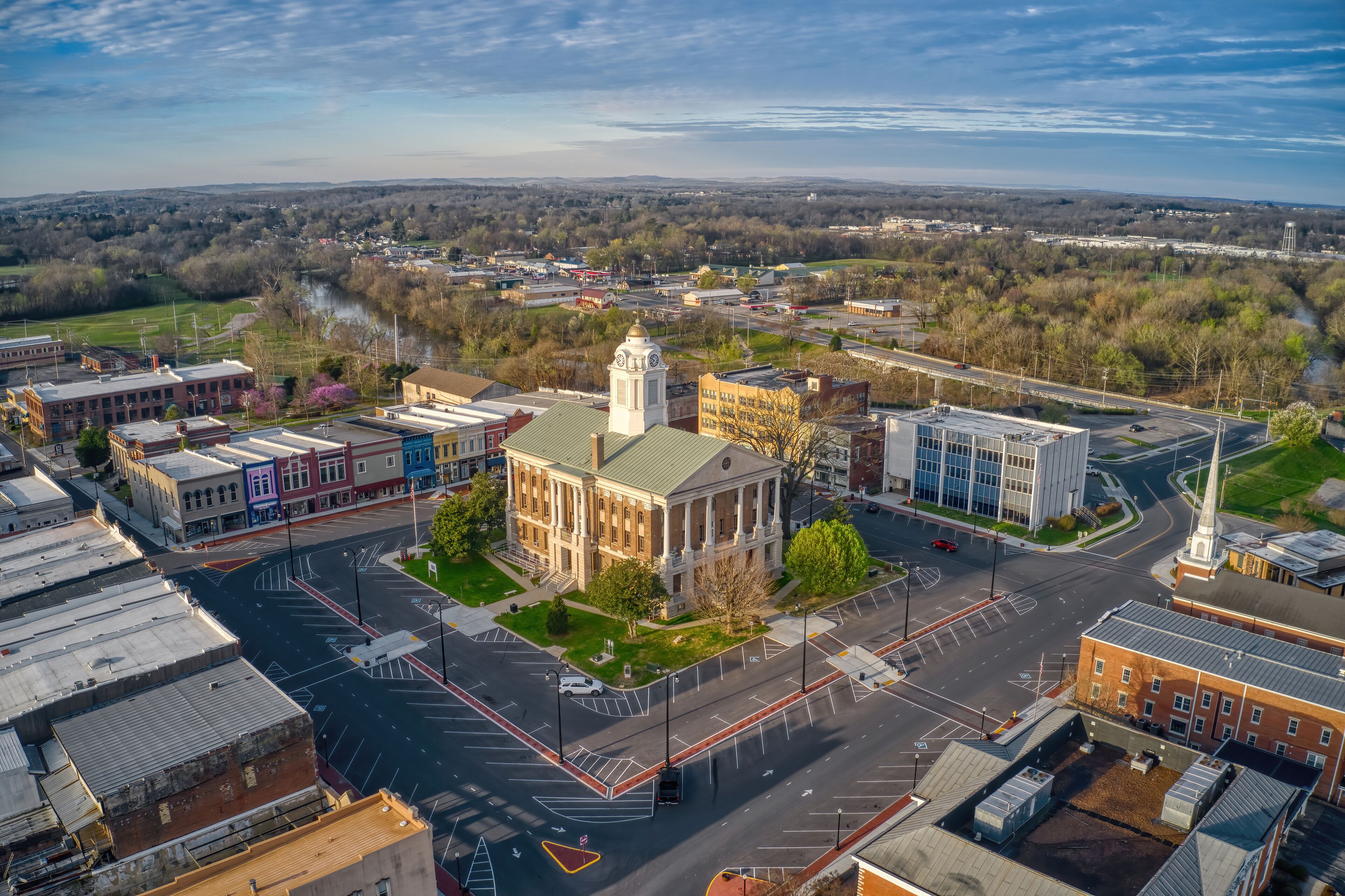 An aerial view of a town with a prominent building with a clock tower, a river, and surrounding greenery.