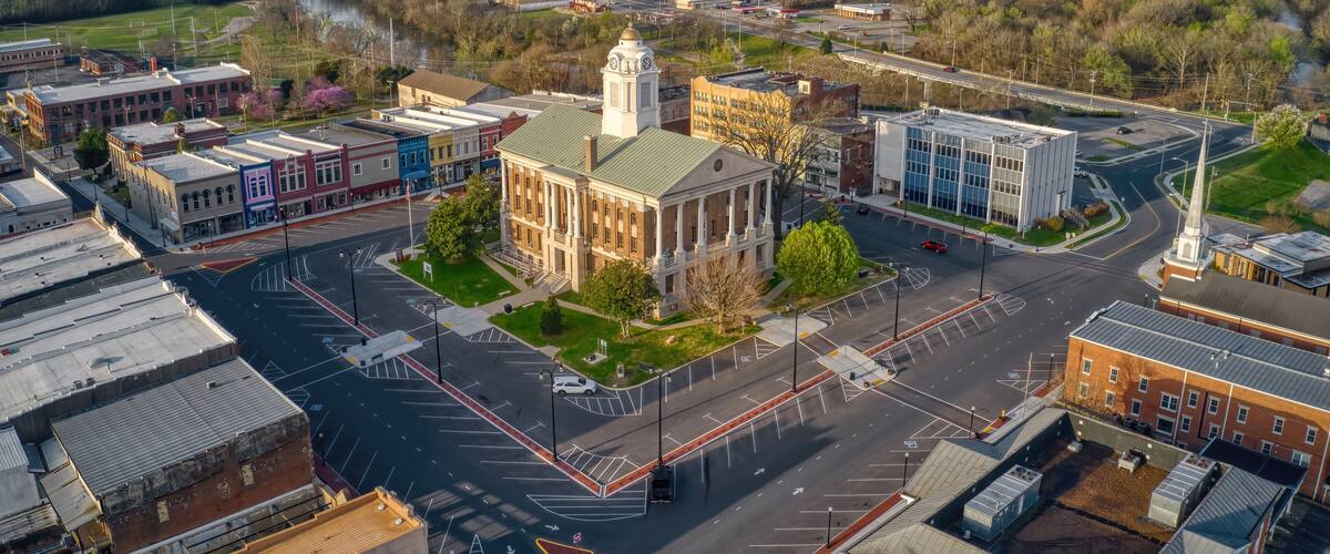 An aerial view of a town with a prominent building with a clock tower, a river, and surrounding greenery.