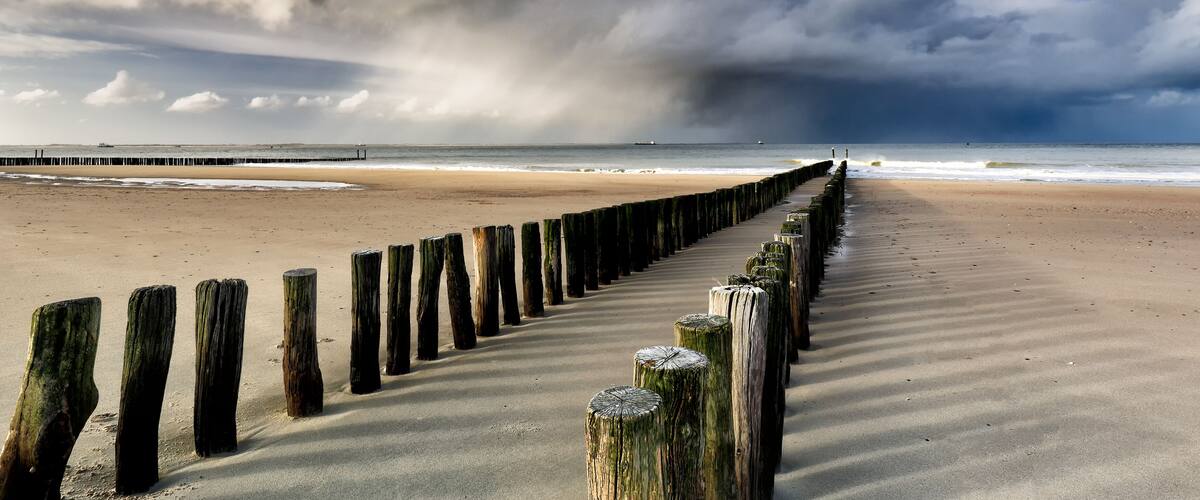 A beach with a wooden jetty extending into the ocean under a cloudy sky.