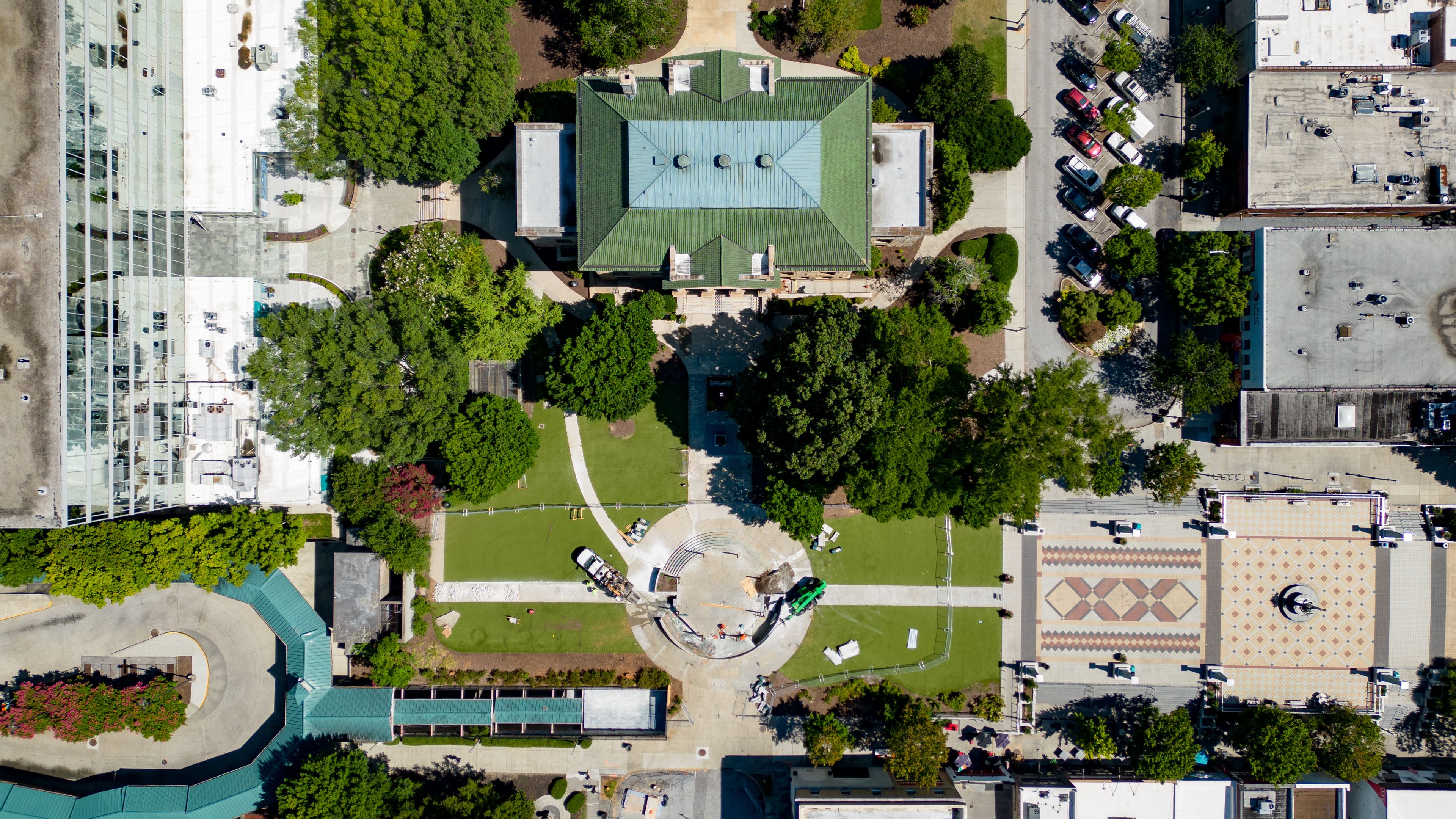 An aerial view of a city with a central green-roofed building surrounded by trees and a circular plaza.