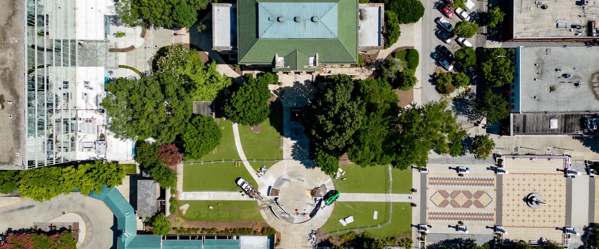 An aerial view of a city with a central green-roofed building surrounded by trees and a circular plaza.